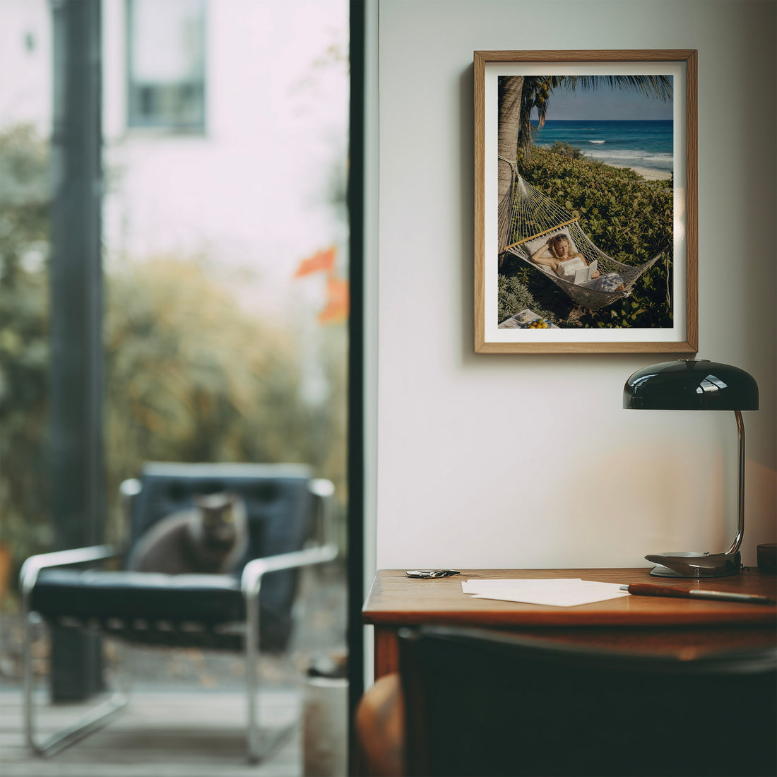 A framed art print depicting a woman relaxing in a hammock by the beach hangs on a light gray wall in a modern office setting. The scene includes a wooden desk with a black lamp, a blurred chair with a cat, and a large window.