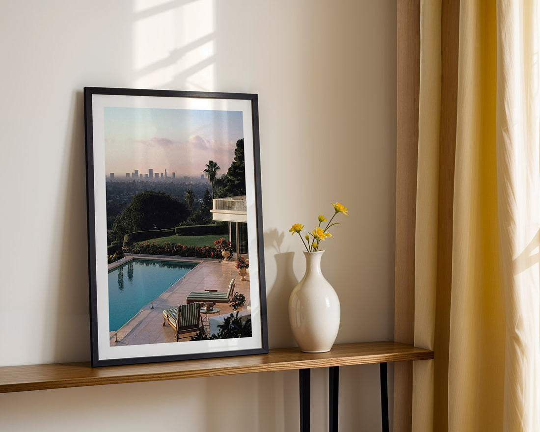 Framed artwork of a pool scene in Los Angeles on a wooden shelf with a vase and flowers.