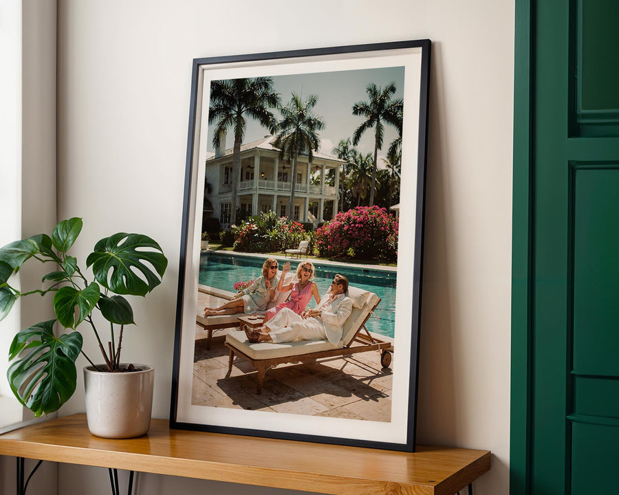 Framed photograph of people by a pool with palm trees, on a wooden shelf with a plant.