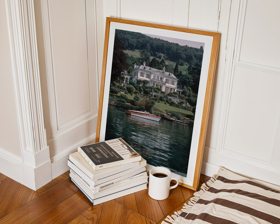 Framed photograph of a house by a lake on a wooden floor with books and a mug.