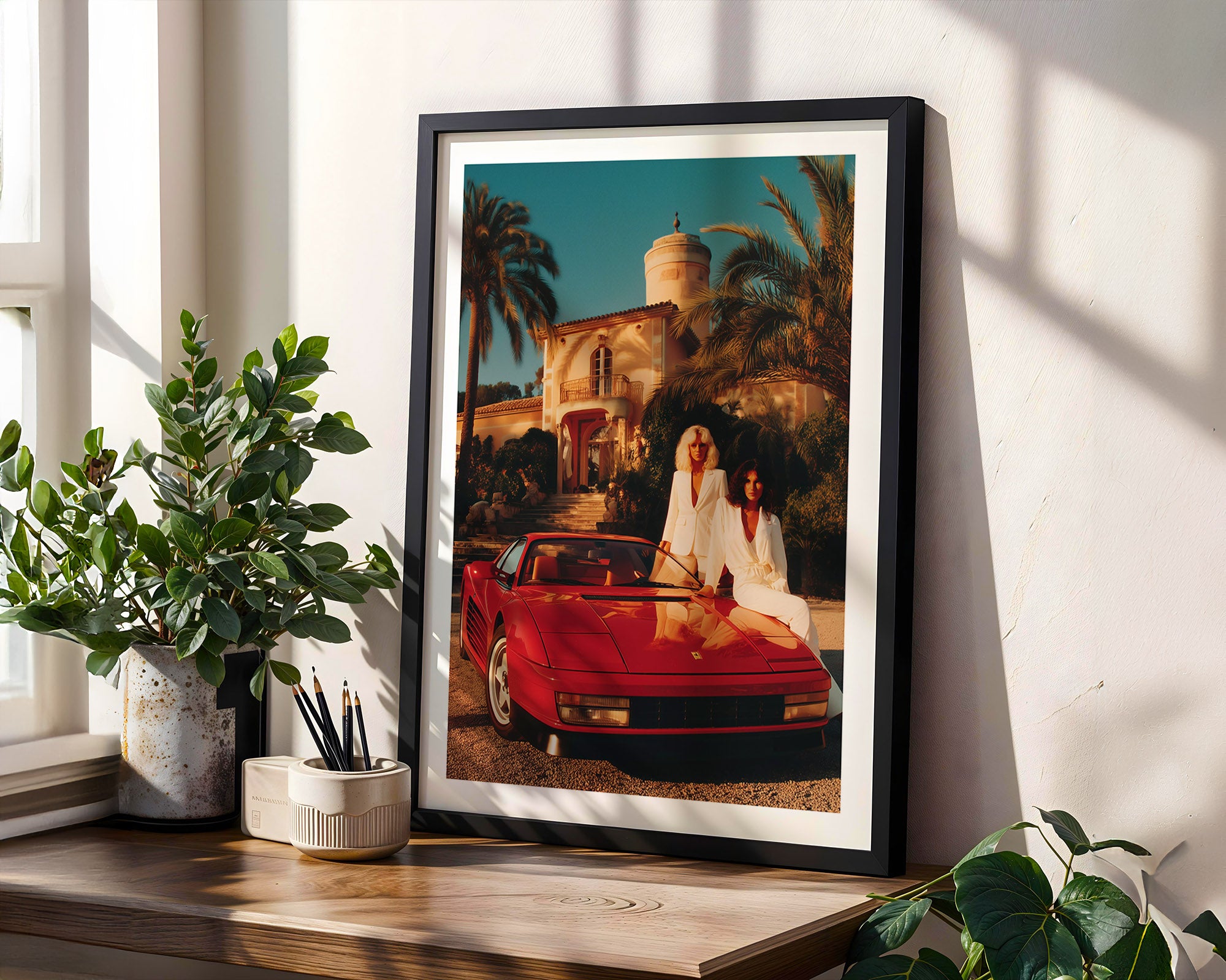 Framed artwork of two people in a red convertible Ferrari Testarossa on a wall with plants and a window in the background.