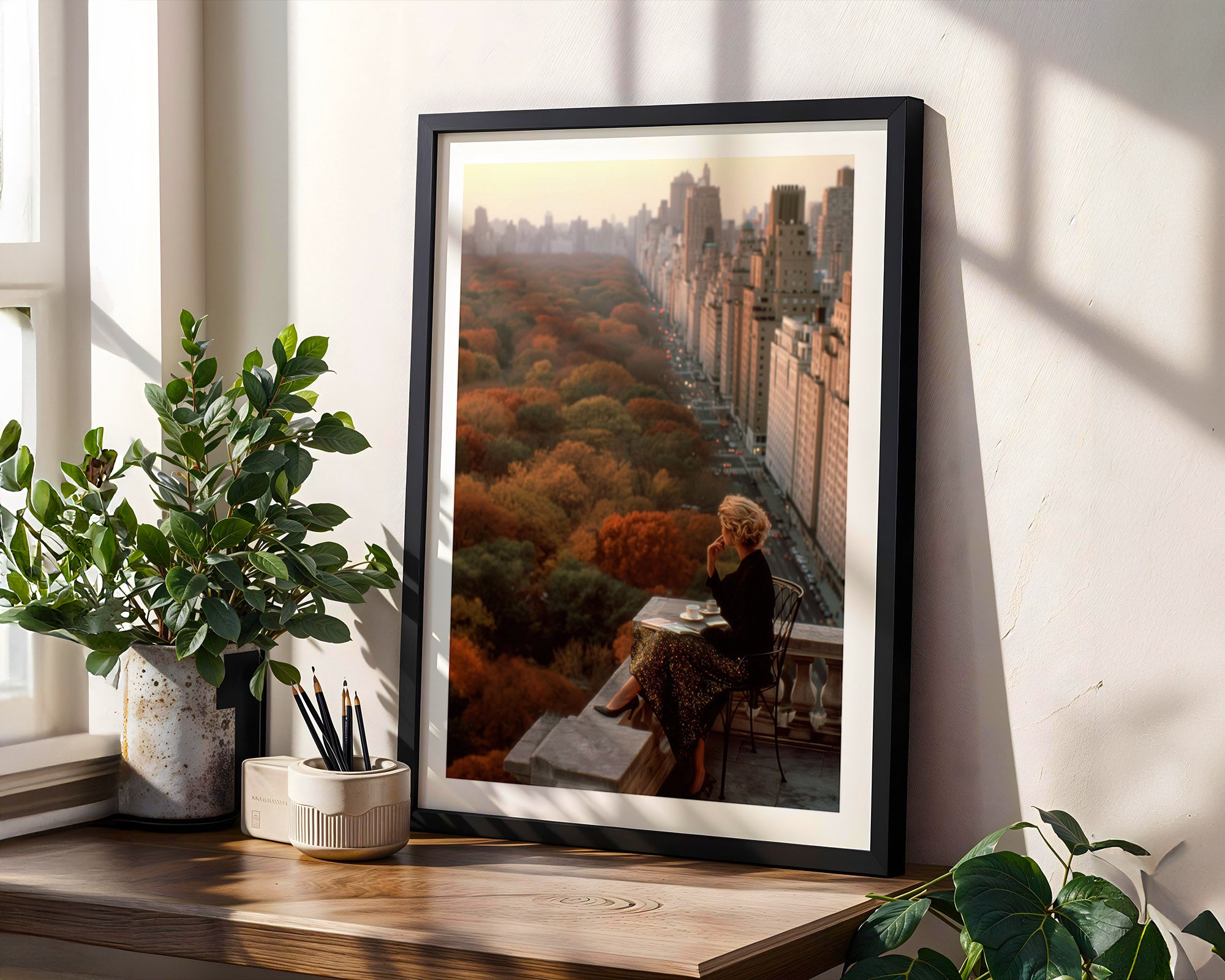 Framed artwork of a cityscape with a person sitting on a edge viewing over Central Park NYC, placed on a wooden surface with plants.