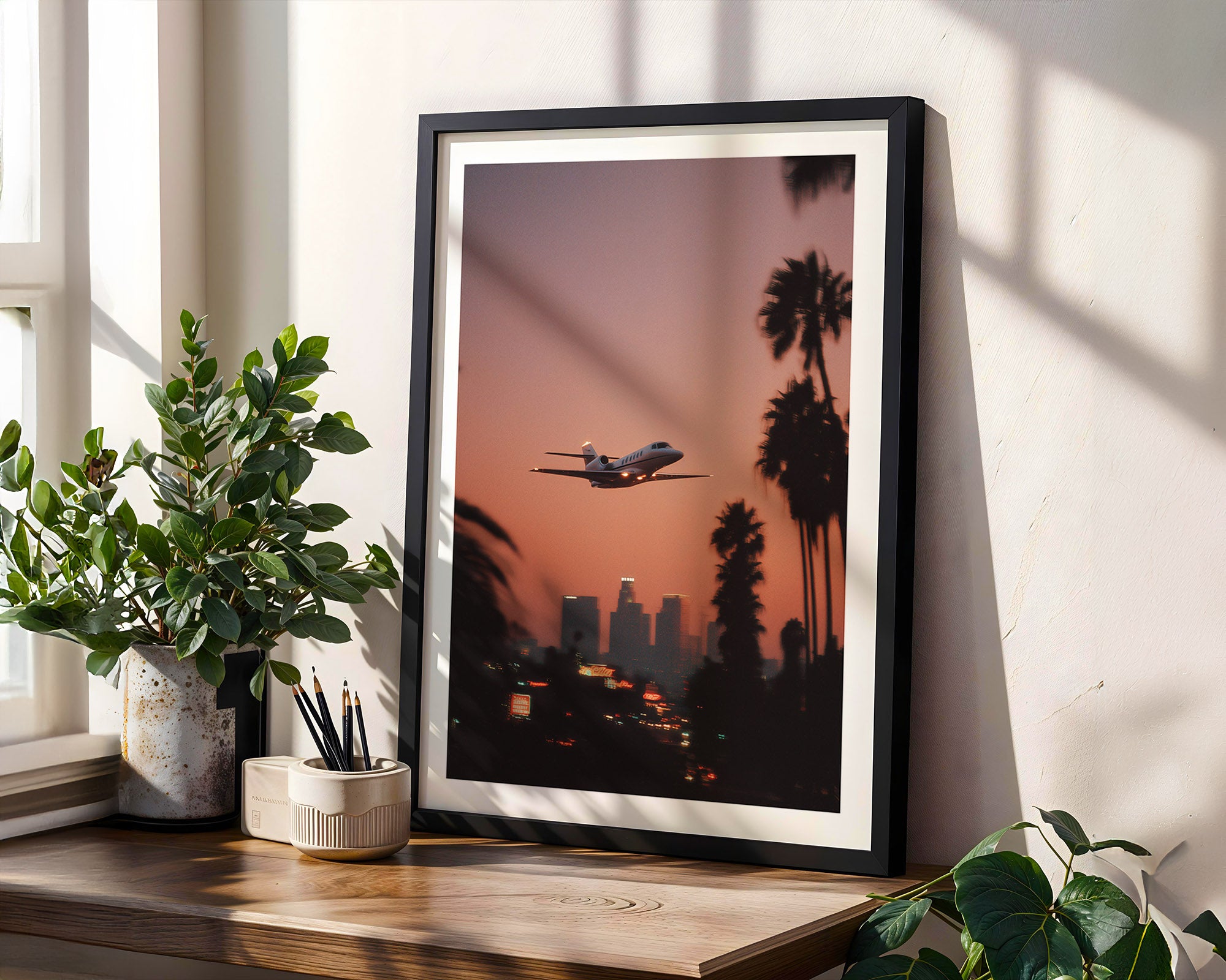 Framed artwork of an airplane flying over Los Angeles with palm trees, placed on a wooden surface with plants.