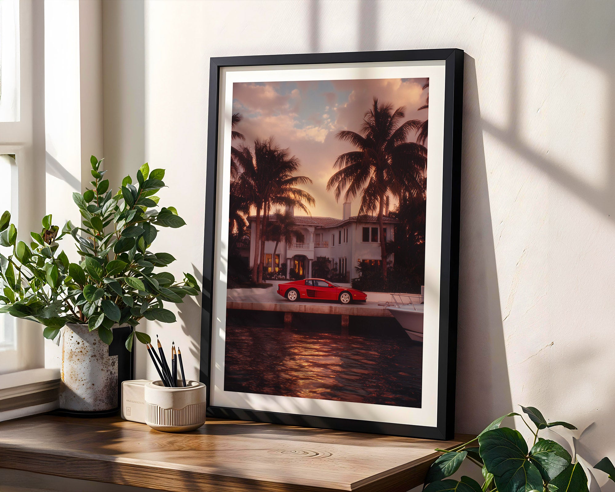 Framed photograph of a red Ferrari Testarossa by palm trees on a wall with plants and a window.