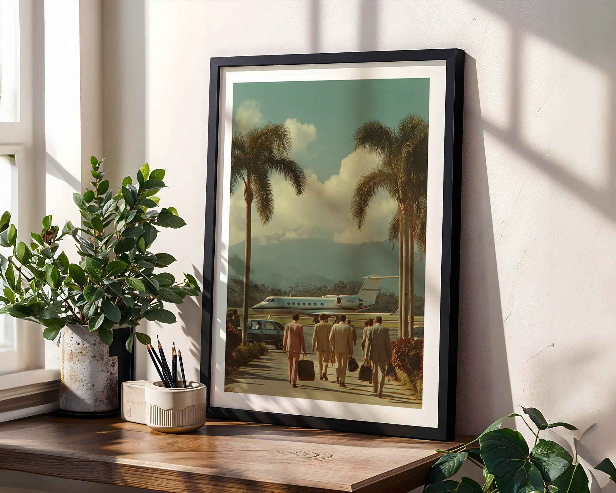 Framed artwork of people walking towards a plane with palm trees in the background, placed on a wooden surface with plants.