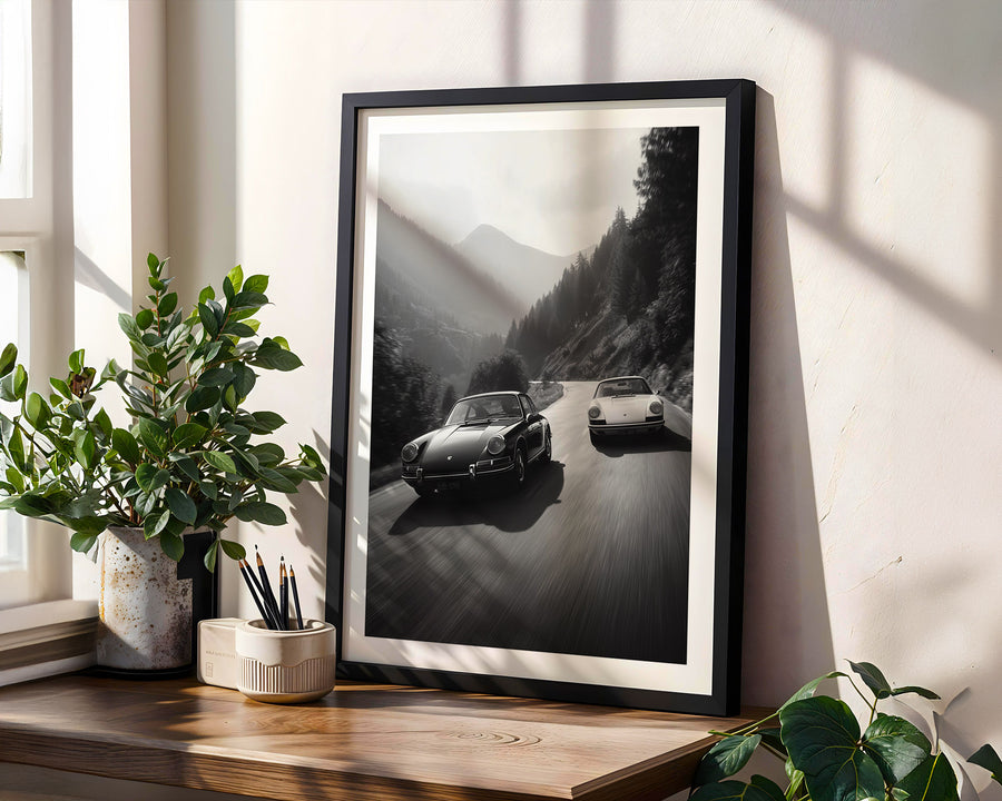 Framed black and white print of two Porsches on a mountain road, placed on a wooden surface with plants.