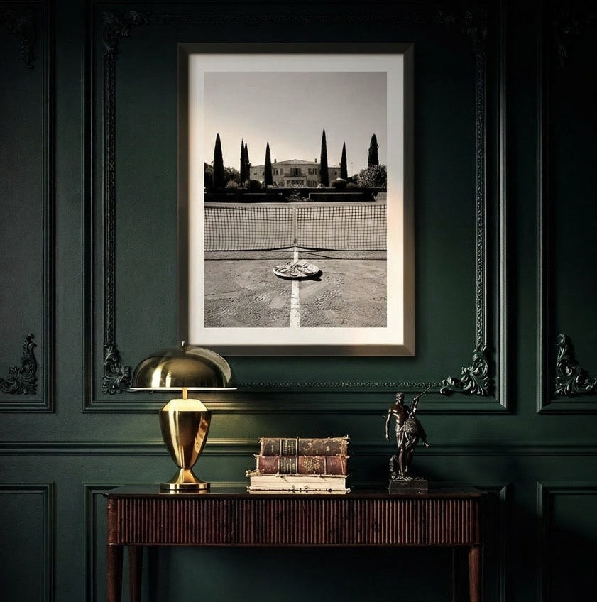 A framed black and white architectural photograph of a classical building and fountain, displayed on a dark green paneled wall above a console table with a gold lamp and books.