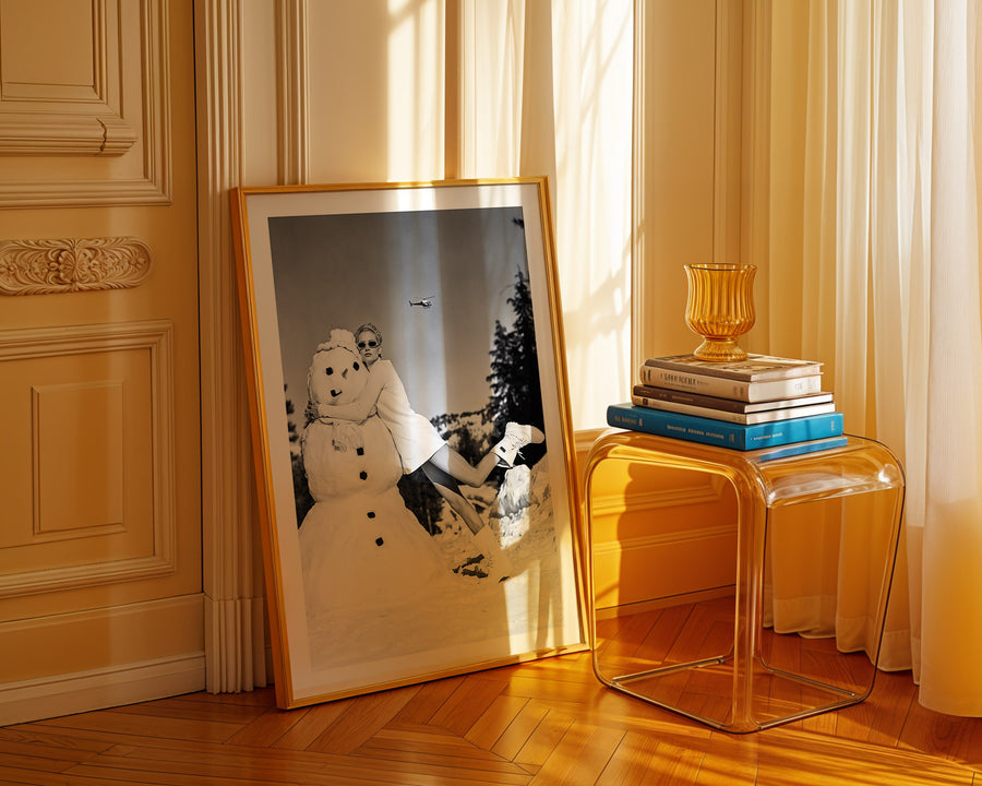 Framed artwork of a snowman on a wooden floor with a clear side table and books in the background.