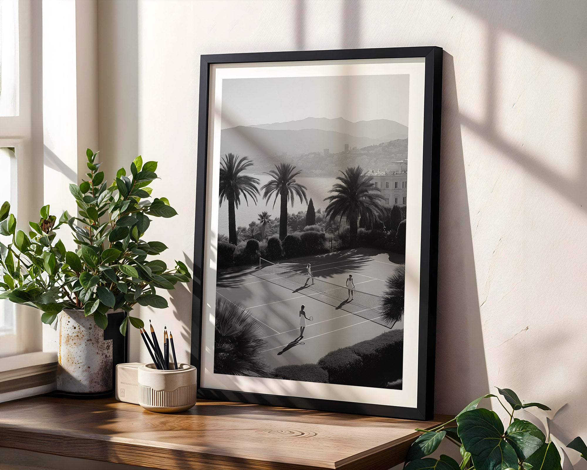 Framed black and white print of a park scene with palm trees on a wall next to a plant.