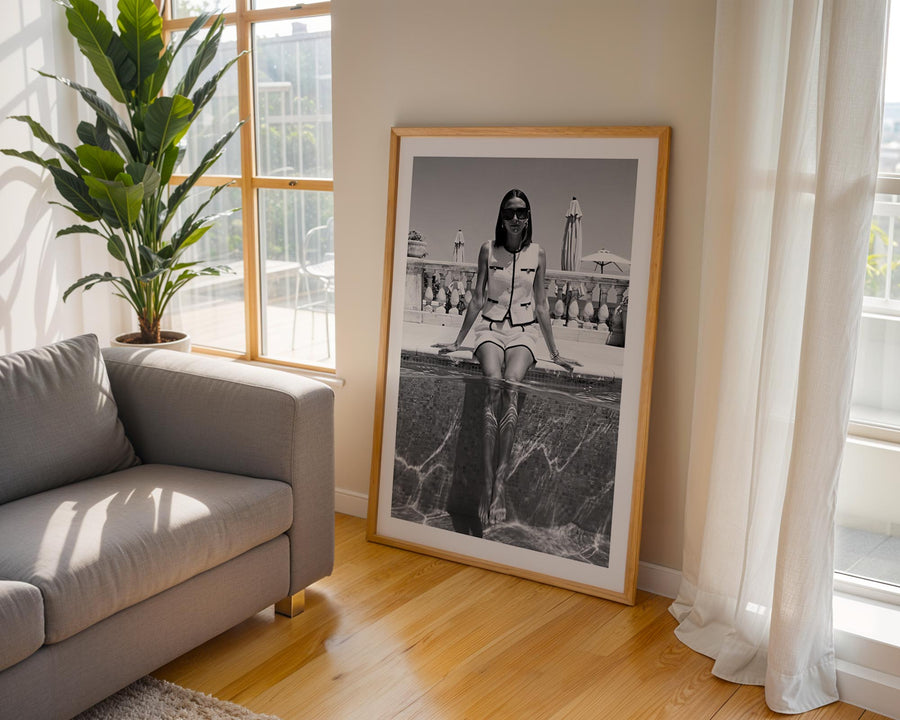 Framed black and white photograph of a woman sitting on a ledge, placed in a room with a gray sofa and large window.