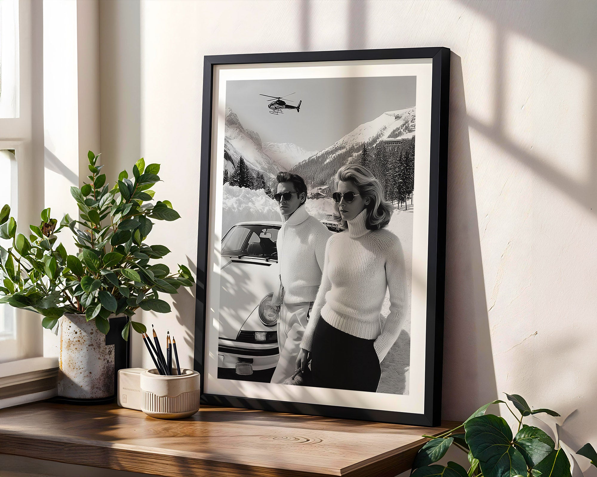 Framed black and white photograph of a couple by a car on a wall with plants around.