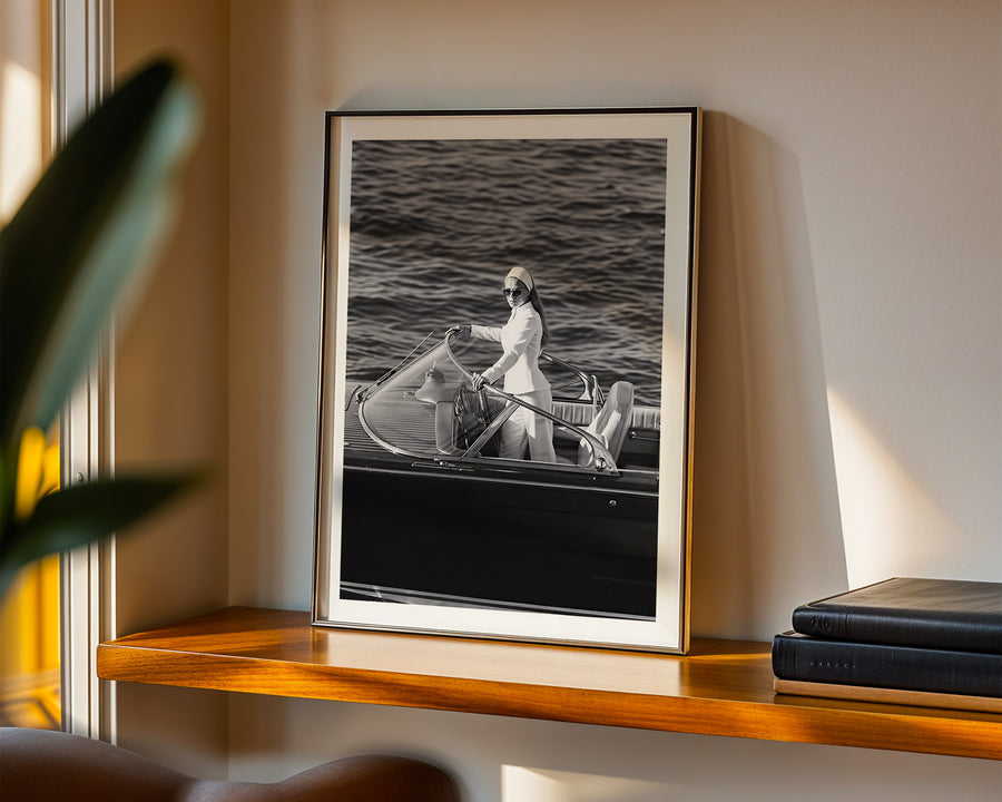 A framed black and white art print of a woman steering a classic motorboat on the water, displayed on a wooden shelf in a warm, modern interior setting.