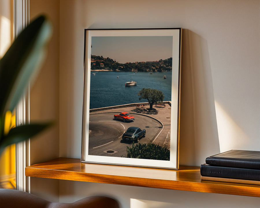 Framed artwork of a coastal scene with cars on a road, displayed on a shelf.