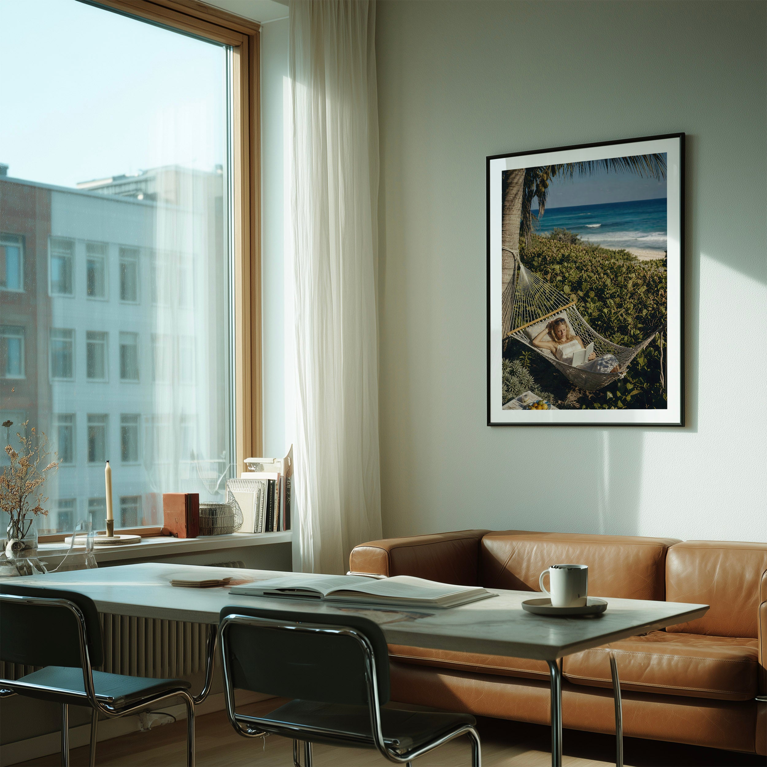 A framed vertical art print depicting a woman relaxing in a hammock on a tropical beach, hanging on a light gray wall in a modern, sunlit living room with a brown leather sofa and a white dining table.