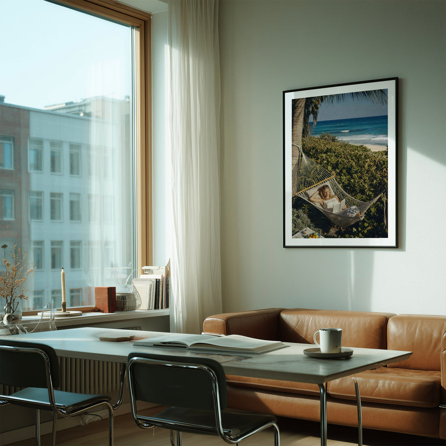 A framed vertical art print depicting a woman relaxing in a hammock on a tropical beach, hanging on a light gray wall in a modern, sunlit living room with a brown leather sofa and a white dining table.