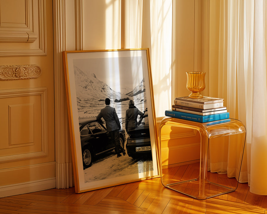 Framed photograph of two people by a car on a wooden floor with a clear side table and books.