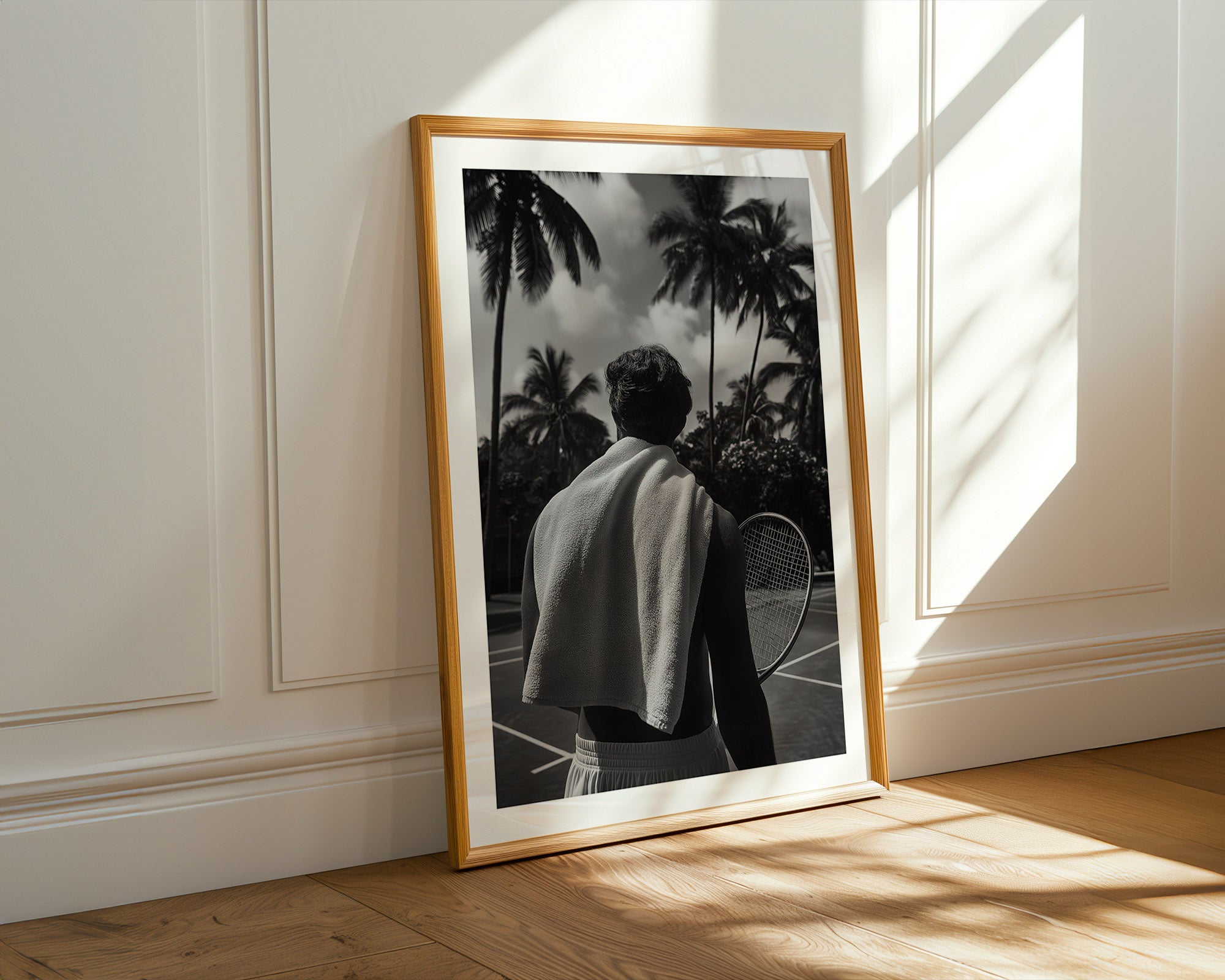 Framed black and white photo of a person holding a tennis racket against a palm tree backdrop, placed on a wooden floor with sunlight casting shadows.