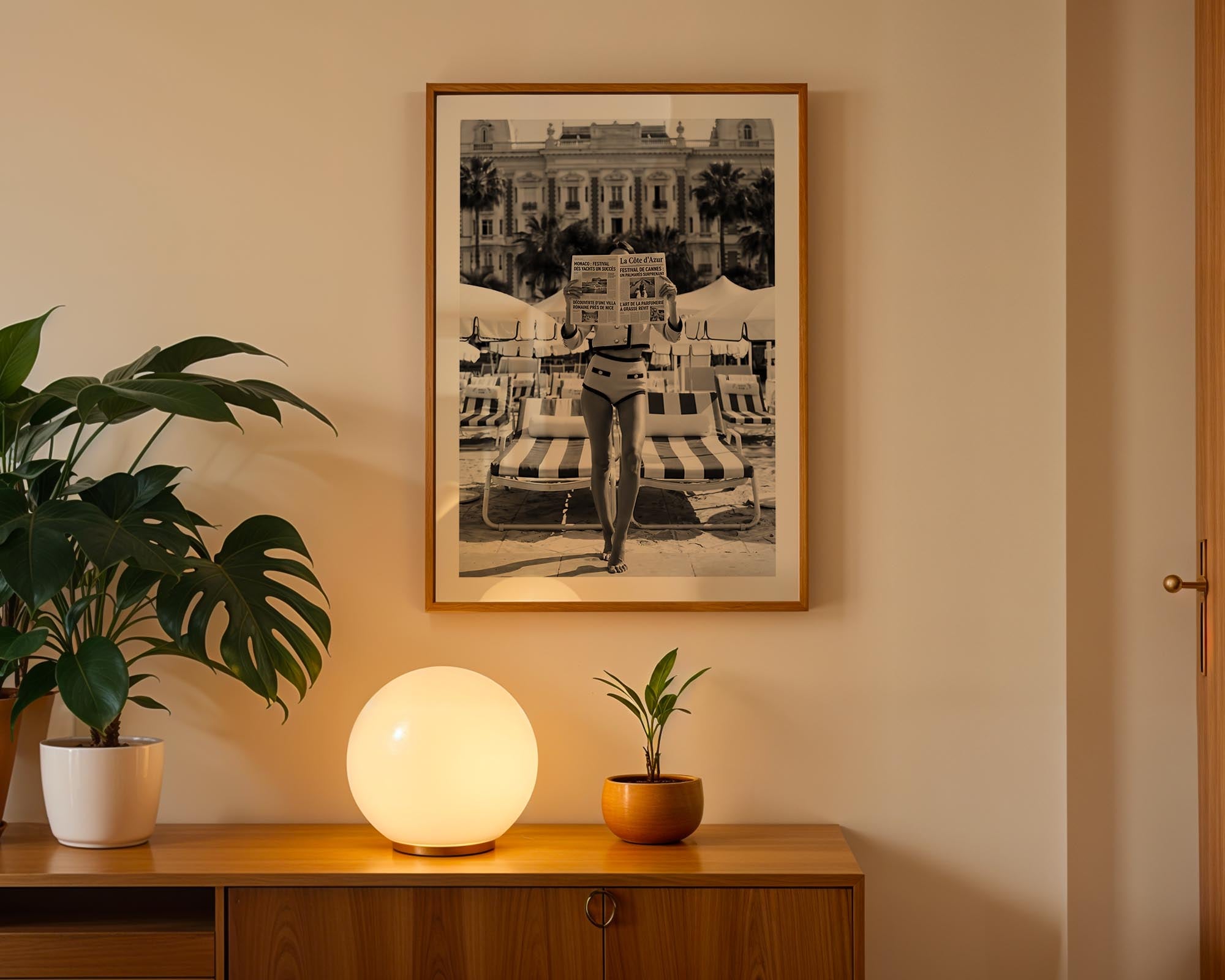 Framed black and white artwork on a wall with a plant and lamp on a wooden surface below.