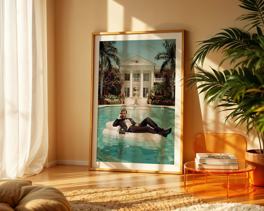 Framed artwork of a man on an inflatable pool raft by a pool, displayed in a room with a plant and table.
