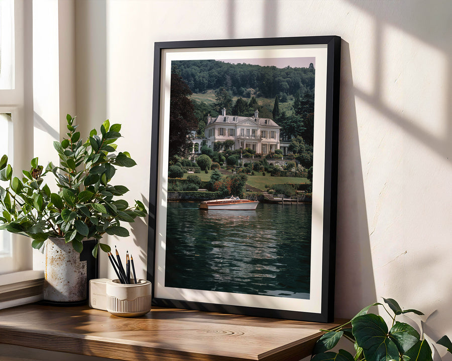 Framed photograph of a house by a lake on a wooden shelf with plants. Lake Lugano.