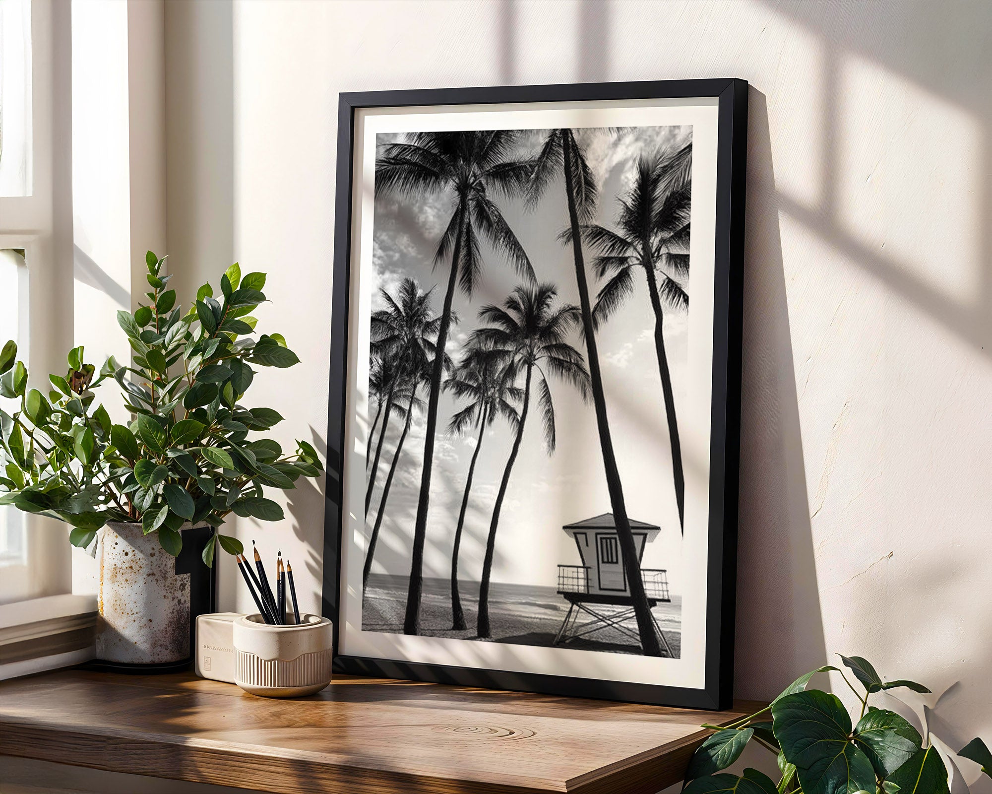 Framed black and white palm tree print on a wall with plants around