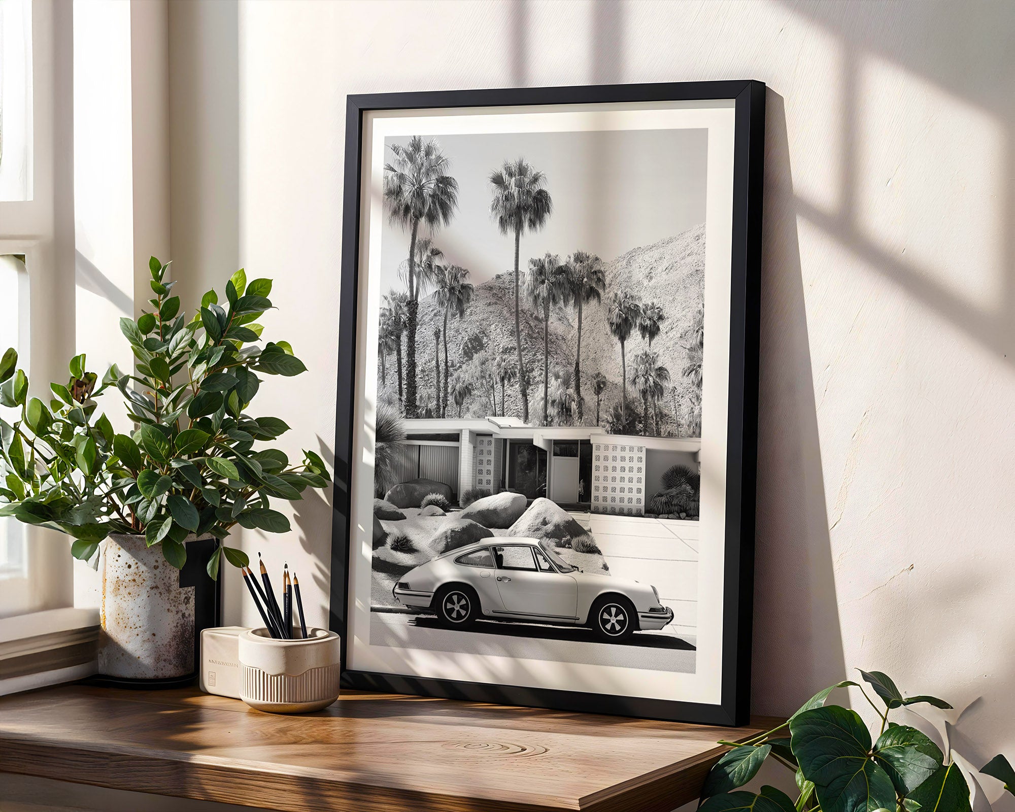 Framed black and white print of a car and palm trees on a wall with plants and decor below.