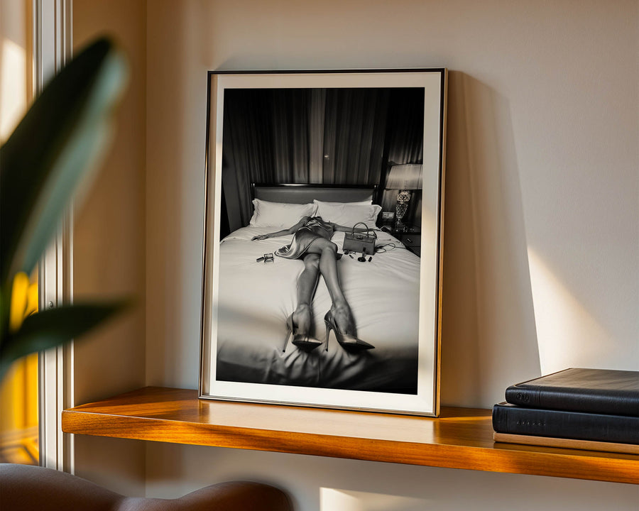 Framed black and white photograph of a person on a bed in a room with a lamp and books.