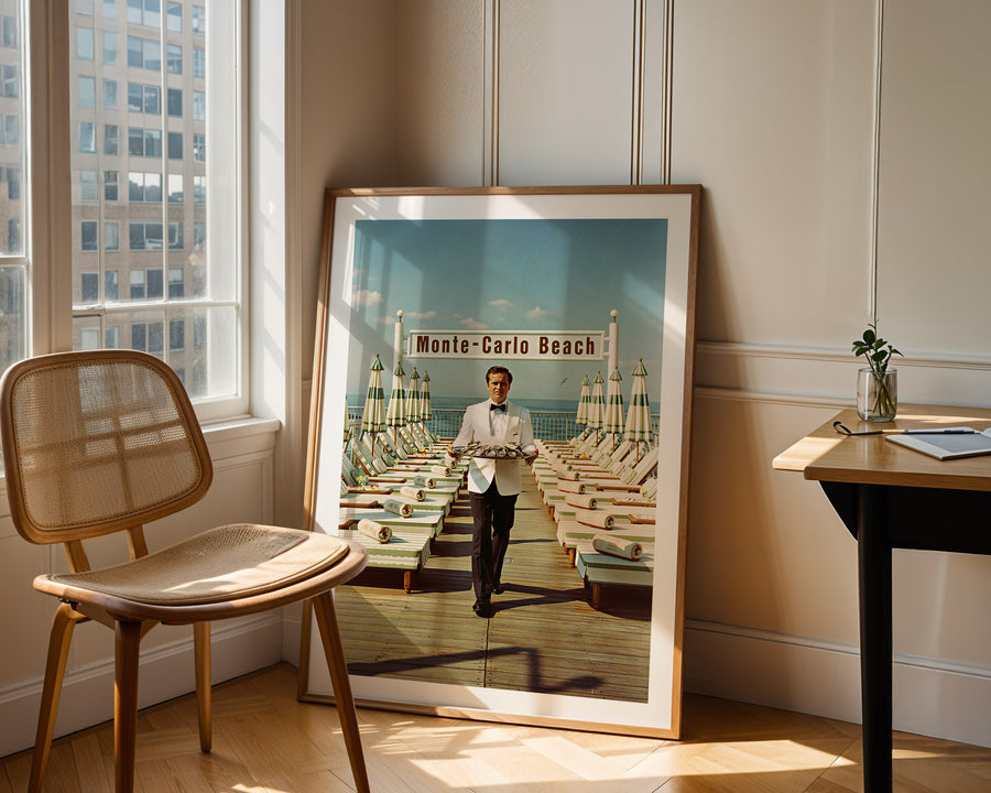 Framed poster of a man in a bow tie walking on a beach with a cityscape in the background, leaning against a wall in a room with a chair and table.
