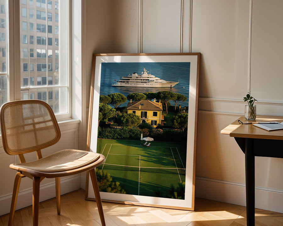 Framed artwork of a yacht and house on a tennis court in a room with a chair and table.