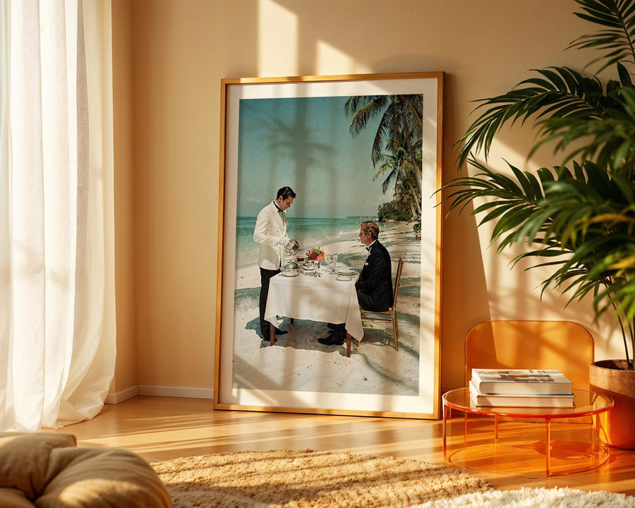 Framed artwork of two people dining on a beach in a living room with a plant and table.