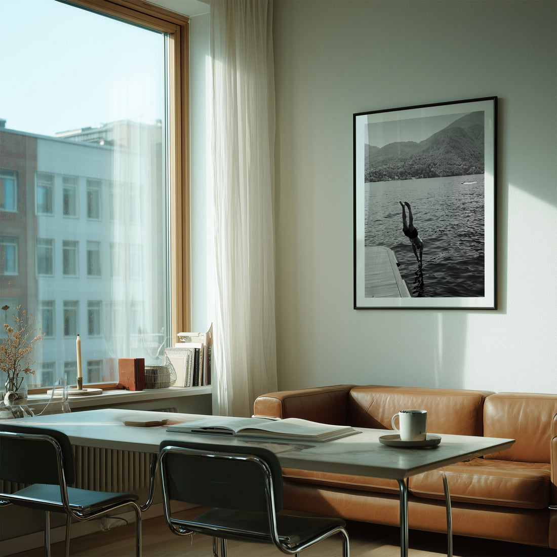 A framed black and white art print of a crane by a lake hangs on a light gray wall in a modern living space with a brown leather sofa, a white dining table, and a large window.