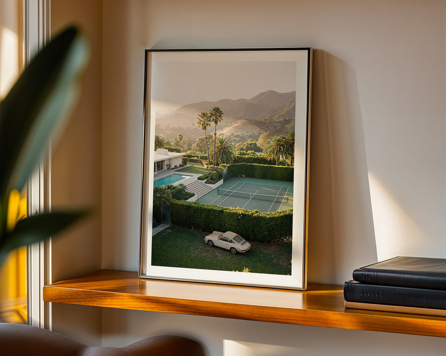 Framed picture of a tennis court with mountains on a wall above a shelf.