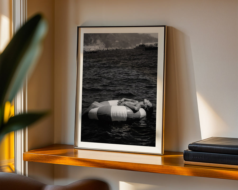 Framed black and white artwork of a person on an inner tube in water, displayed on a wooden shelf.