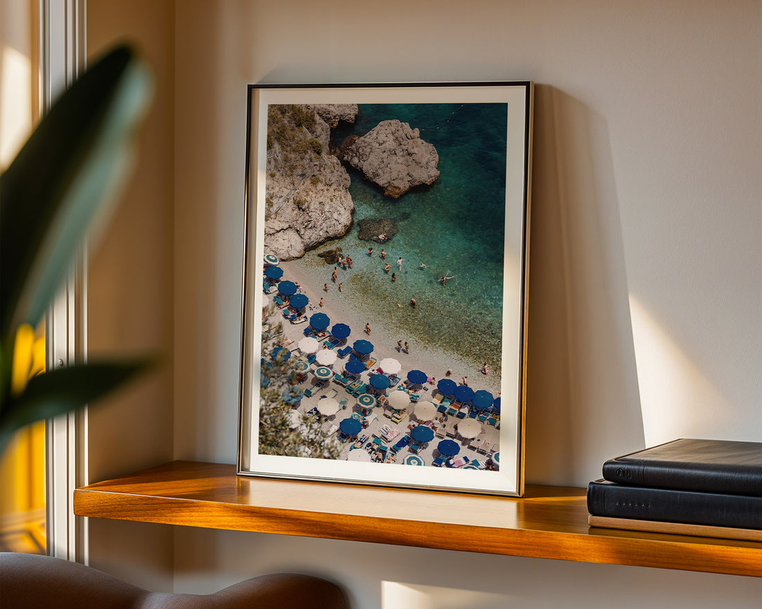 Framed artwork of a beach scene with blue umbrellas on a wooden shelf.