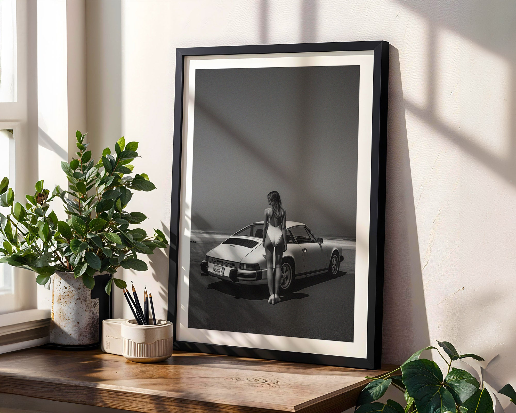 Framed black and white photo of a woman standing next to a car on a wooden shelf with plants.