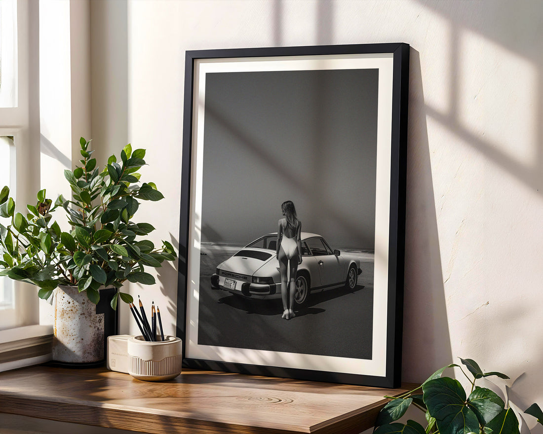 Framed black and white photo of a woman standing next to a car on a wooden shelf with plants.