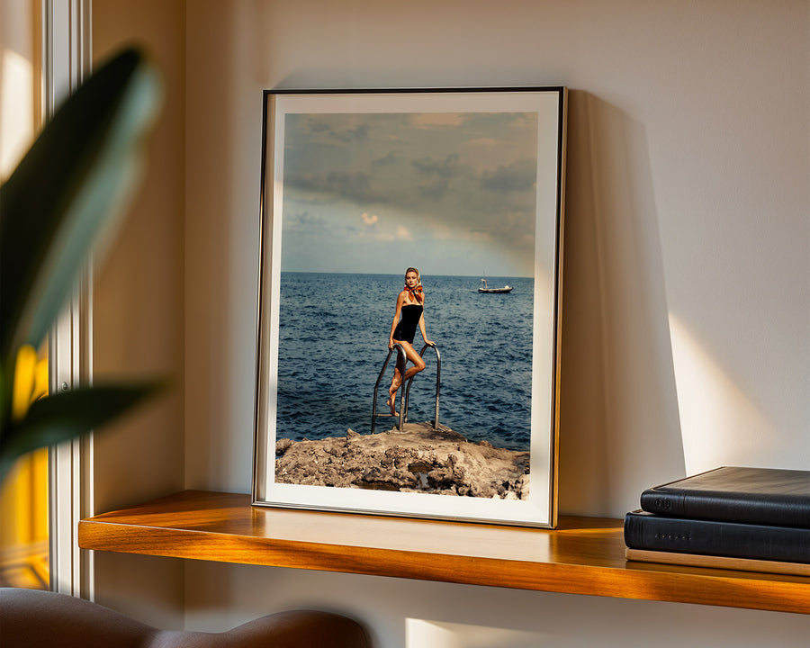 A framed art print featuring a vintage photograph of a woman in a black swimsuit riding a penny-farthing bicycle on a rocky shoreline, displayed on a wooden shelf in a warmly lit room.