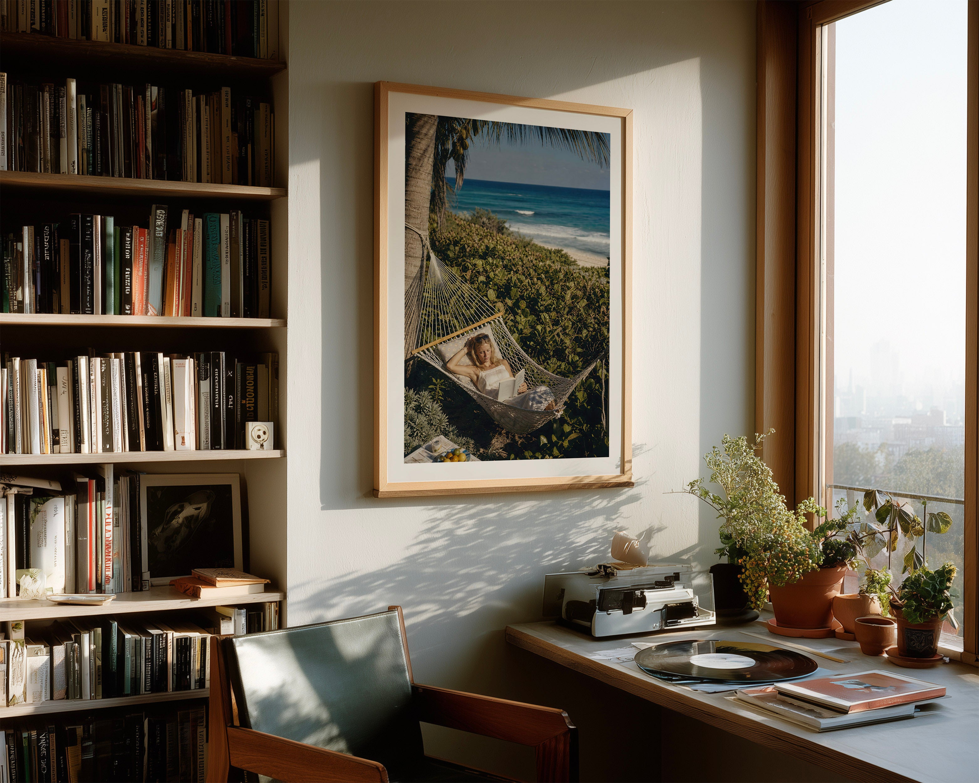 A framed art print depicting a woman relaxing in a hammock on a tropical beach, hanging on a wall in a sunlit home office next to a bookshelf and desk.