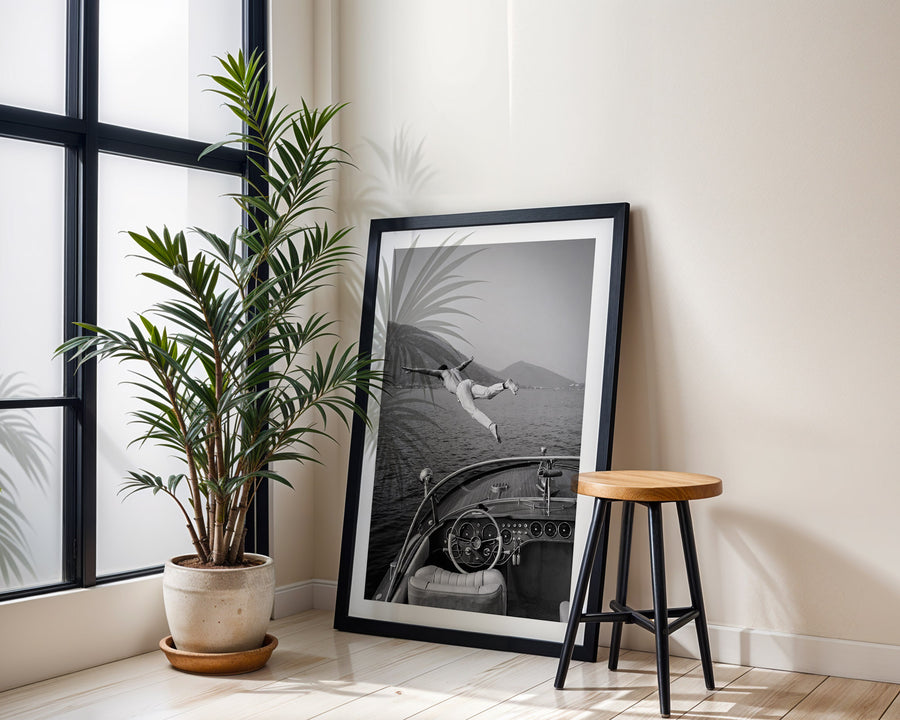 Framed black and white artwork of a vintage car interior next to a potted plant and wooden stool.