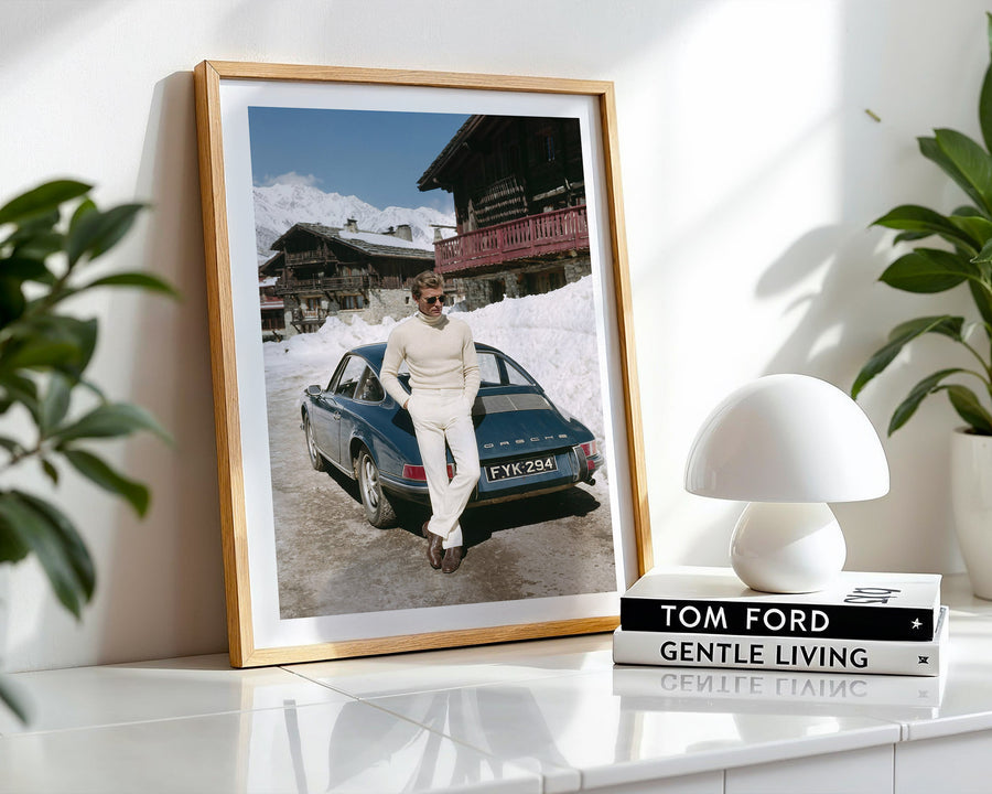 Framed photograph of a person next to a vintage car on a snowy mountain road, placed on a white surface with a lamp and books.