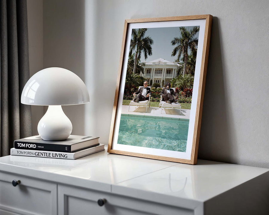 Framed photograph of a pool scene on a white surface with a lamp and books.