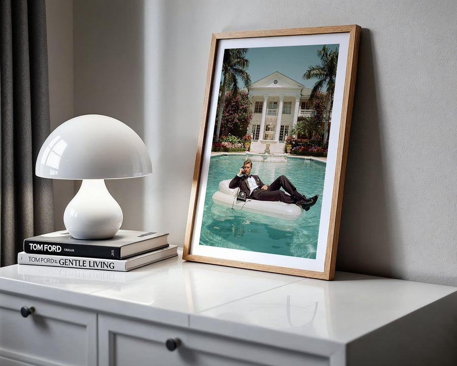Framed photograph of a person on an inflatable raft in a pool, placed on a white surface with a lamp and books.