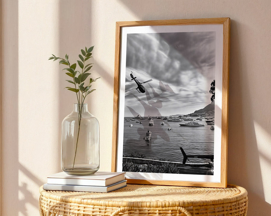 Framed black and white artwork of a lake scene with boats and a helicopter, placed on a wicker side table with a vase and books.