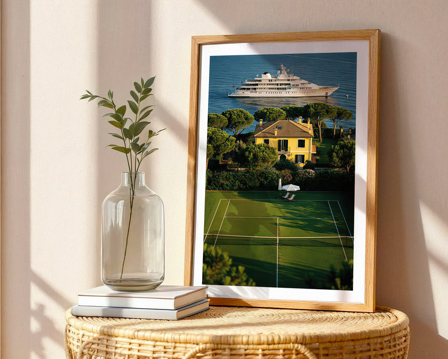 Framed artwork of a house with a tennis court and ship on a wall, next to a vase and books on a table.