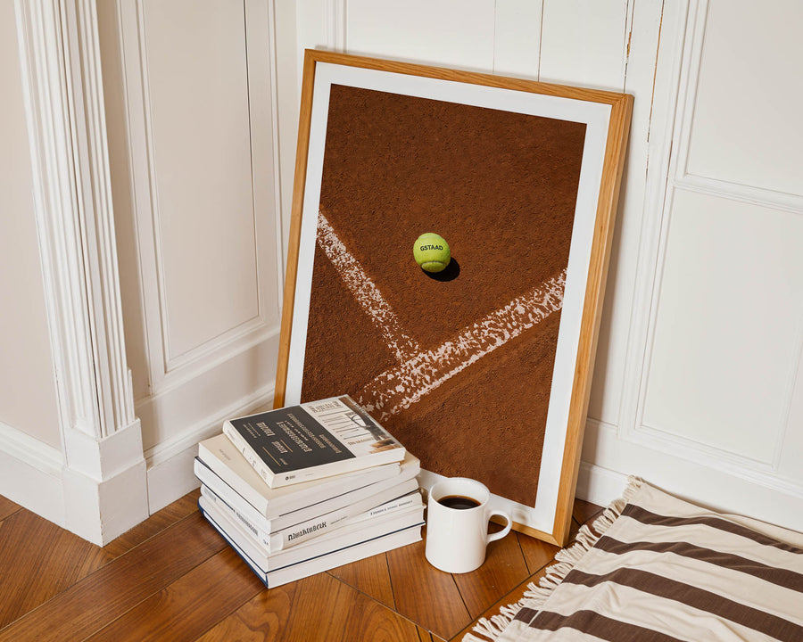 Framed artwork of a tennis ball on a brown background next to books and a mug on a wooden floor.