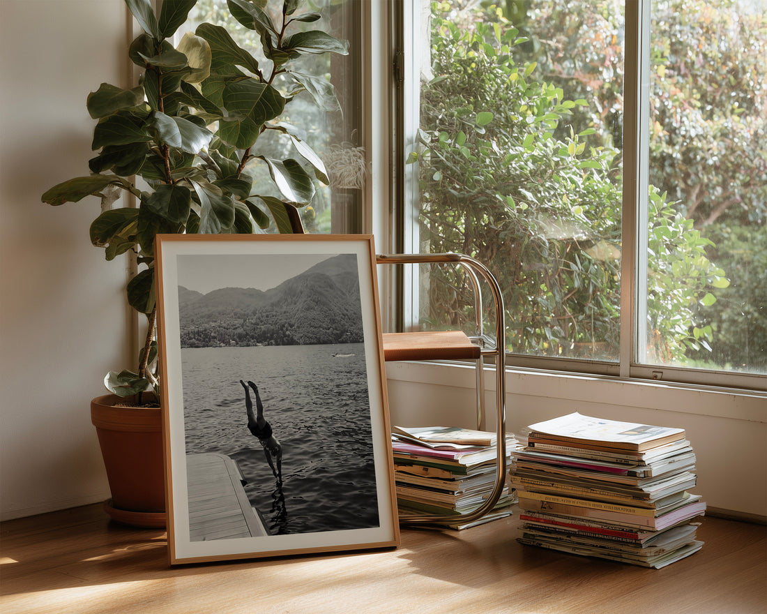 A framed black and white art print depicting a swan on a dock by a lake, styled on a wooden floor next to a potted fiddle-leaf fig plant and a brass side table holding stacks of magazines in a sunlit room.