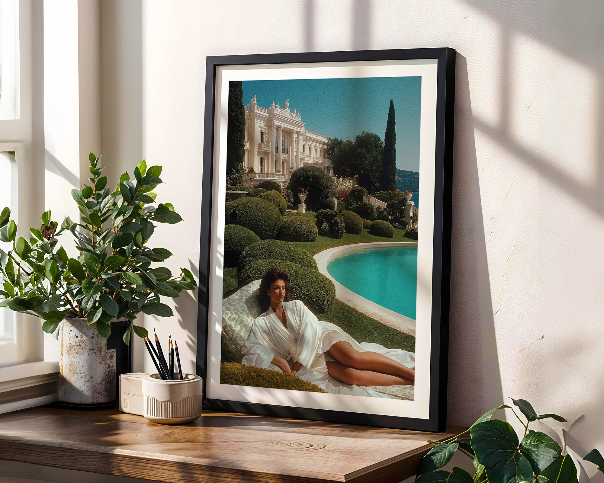 Framed artwork of a woman in a white robe by a pool with a classical building in the background, on a wooden shelf with plants.