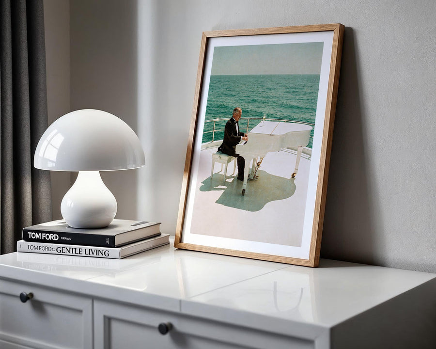 Framed artwork of a man playing a piano on a boat, placed on a white surface with a lamp and books.