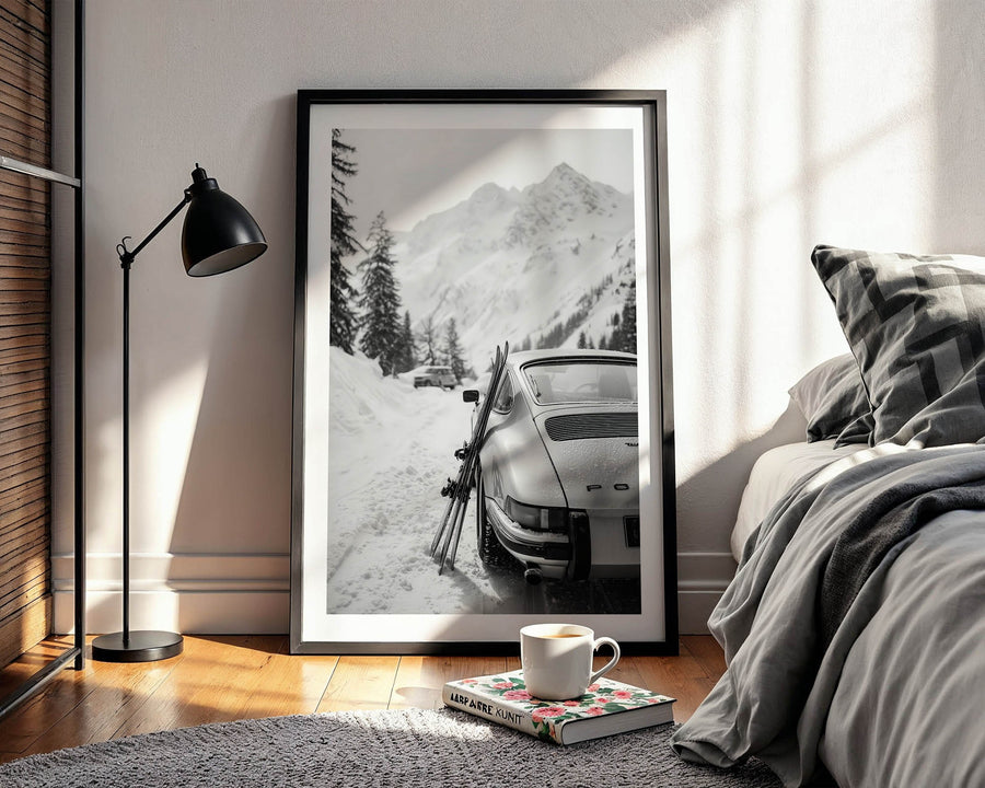 Framed black and white print of a snowy mountain scene with a porsche 911, placed on a wooden floor in a room with a bed and lamp.