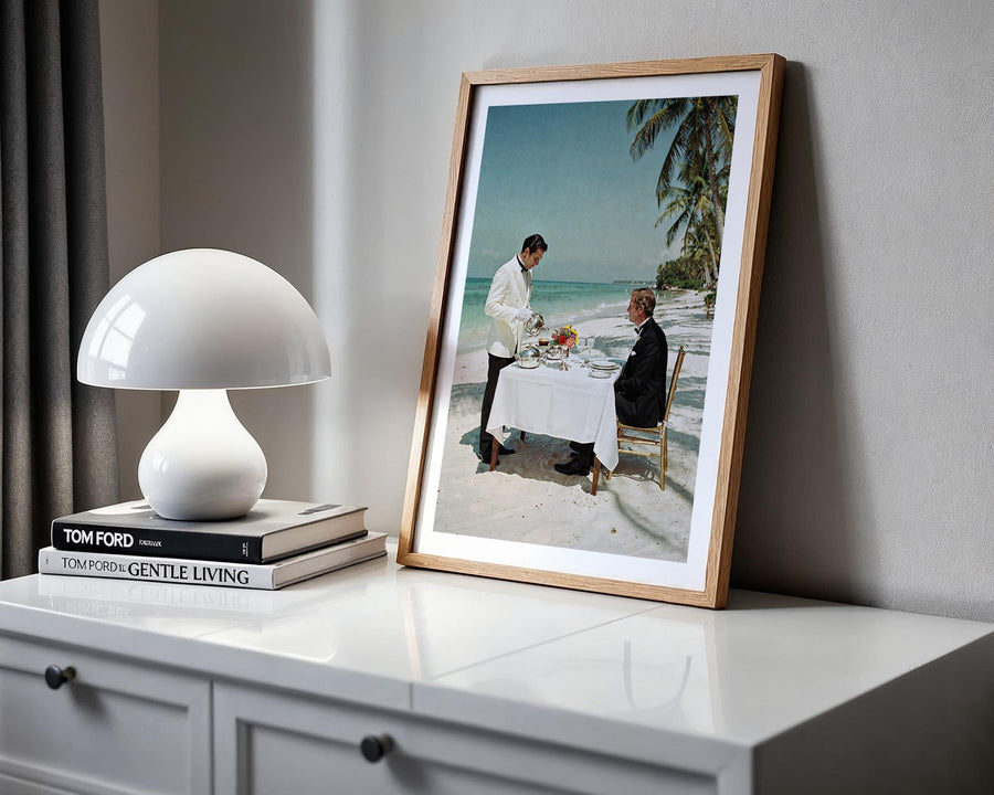 Framed photograph of a couple on a beach next to a white lamp and books on a shelf.